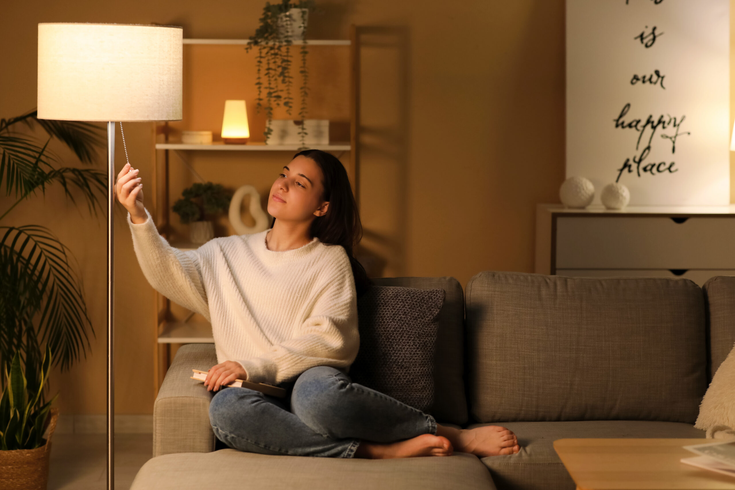 A young woman sitting on a couch turns on a floor lamp to her right.